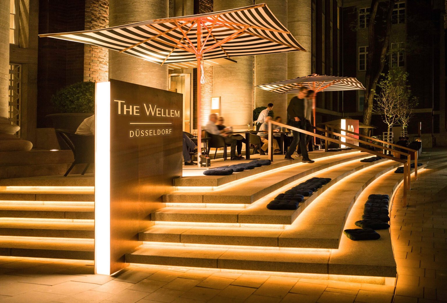 Abendliche Außenaufnahme des Hotel The Wellem in Düsseldorf mit elegant beleuchteter Eingangstreppe, modernen Sonnenschirmen und stilvoller Terrasse.