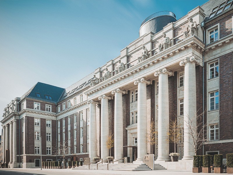 Tagesaufnahme der beeindruckenden historischen Säulenfassade des Luxushotels The Wellem in Düsseldorf mit klassizistischen Figuren und großzügiger Freitreppe unter klarem Himmel.