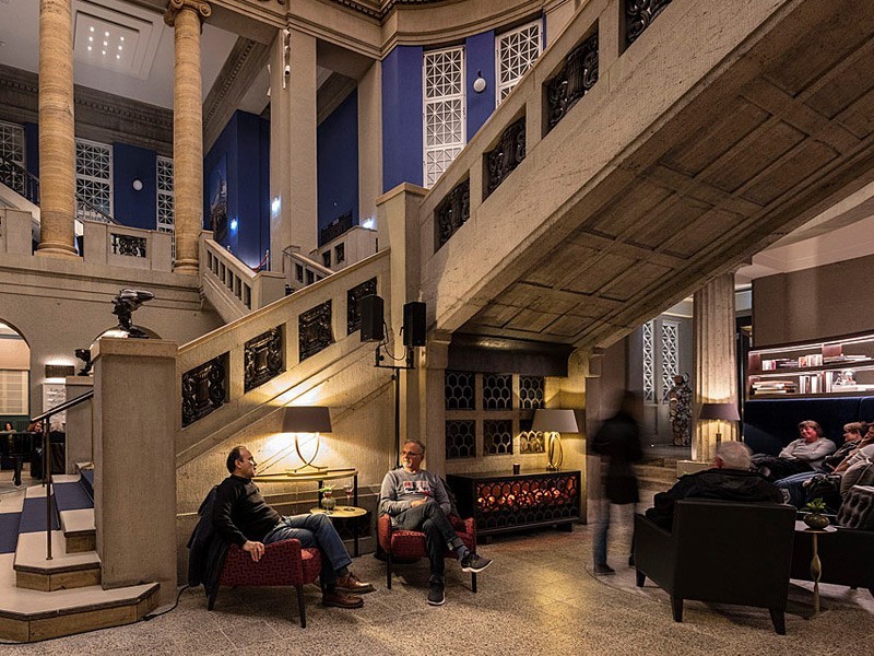 Elegante Lounge unter der historischen Treppe im Hotel The Wellem Düsseldorf mit gemütlichen Sitzbereichen, stimmungsvoller Beleuchtung und klassizistischer Architektur.