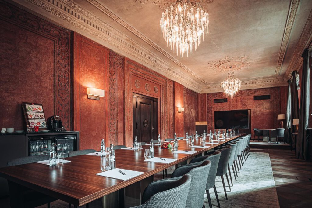 Historischer Salon im Hotel The Wellem in Düsseldorf, ehemals Richterzimmer, mit prachtvollen Stuckdecken, Kronleuchtern, roten Wandvertäfelungen und elegant gedecktem Konferenztisch für exklusive Meetings und Events.
