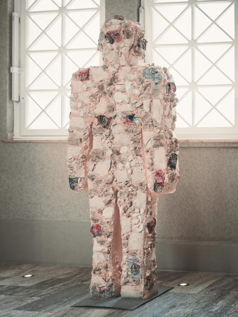 Zeitgenössische Figurenskulptur im Hotel The Wellem Düsseldorf, gefertigt aus strukturierten Fragmenten und eingearbeiteten Dosenreliefs, präsentiert vor großen geometrischen Fensterflächen.