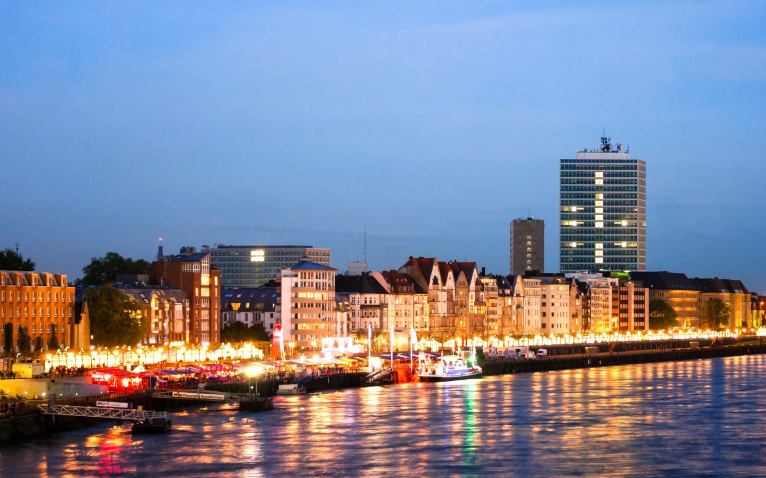Beleuchtete Rheinuferpromenade in Düsseldorf bei Abenddämmerung mit historischen Fassaden, Rheinblick und reflektierenden Lichtern auf dem Wasser am Hotel The Wellem