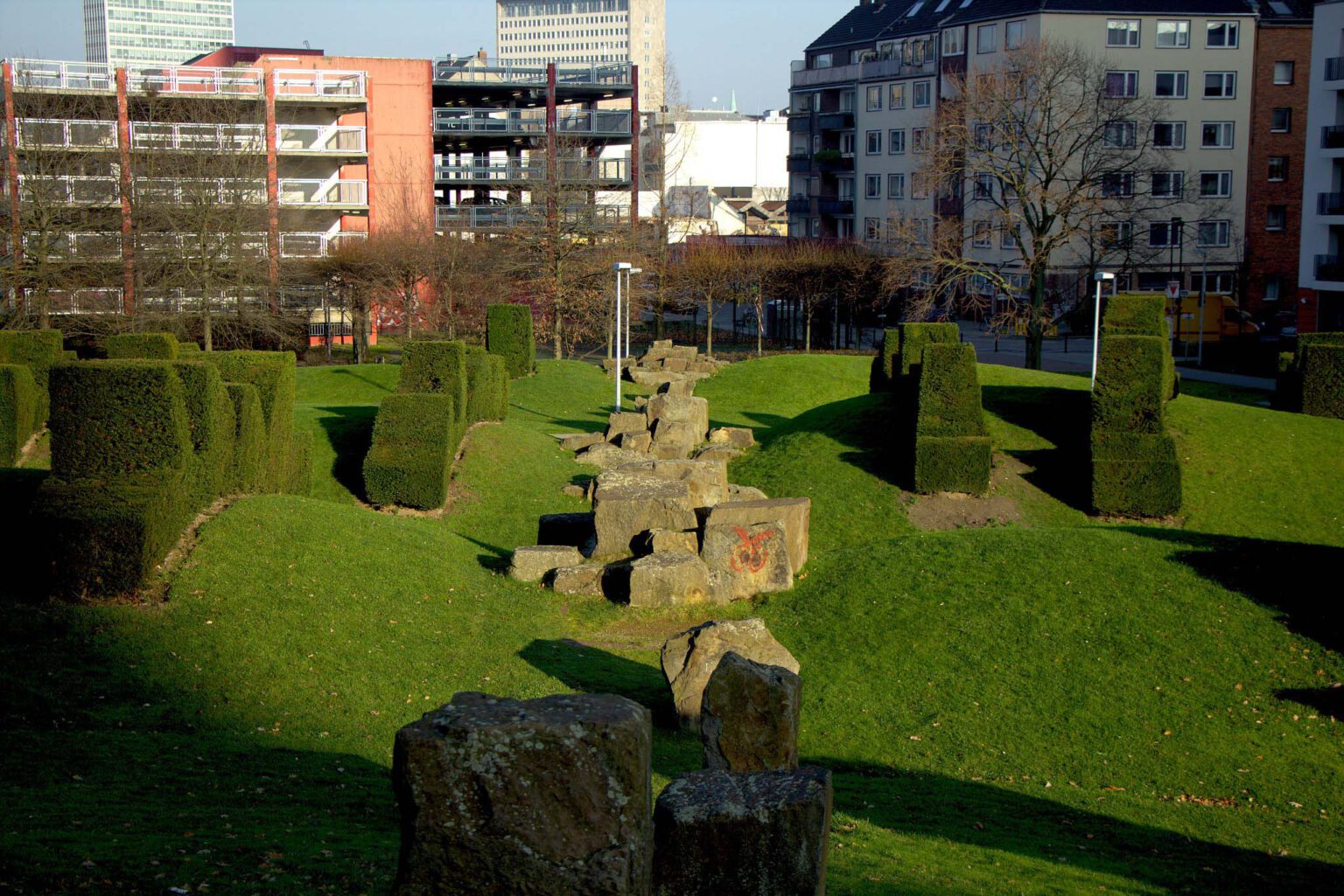 Stein–Installation im Park in Düsseldorfer mit geometrisch geschnittenen Hecken und umliegender urbaner Architektur.
