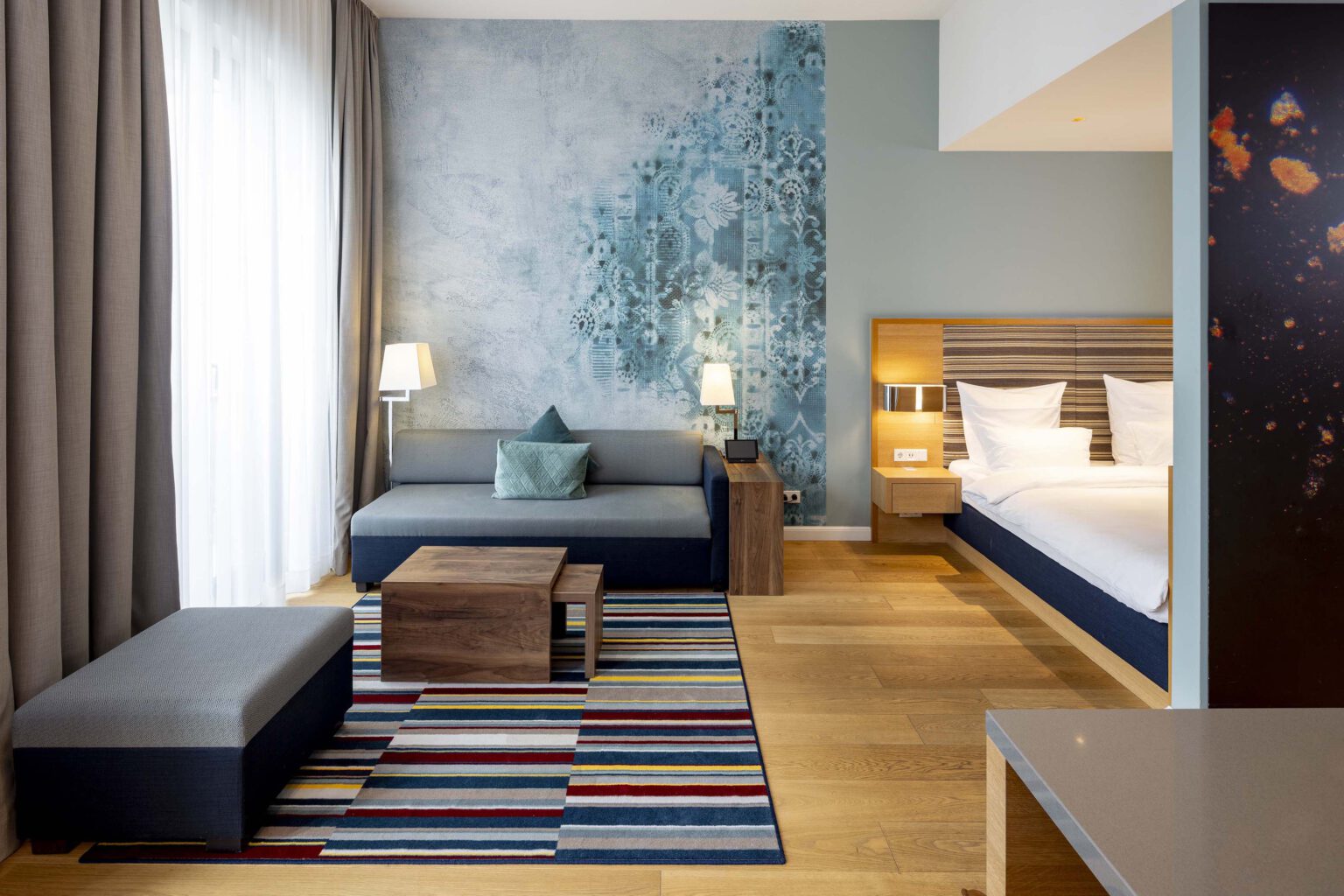 Großzügige Business Suite im Hotel The Wellem Düsseldorf mit modernem Wohnbereich, farbiger Akzentwand, stilvollem Sofa, Designteppich und offenem Schlafbereich.