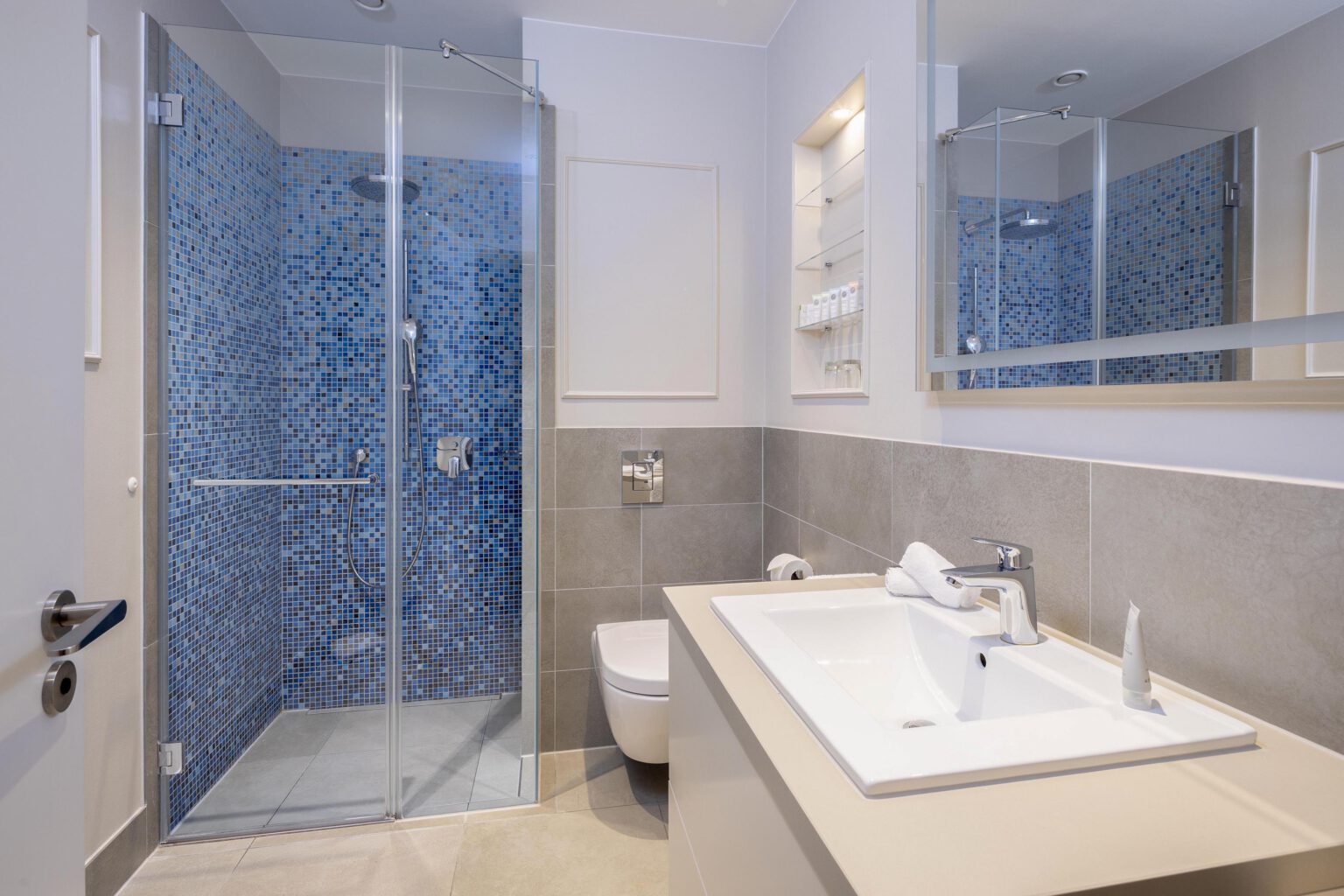 Modern bathroom in the Executive Suite at Hotel The Wellem Düsseldorf with walk-in shower in blue mosaic look, large washbasin, light-colored tiles and high-quality care products.
