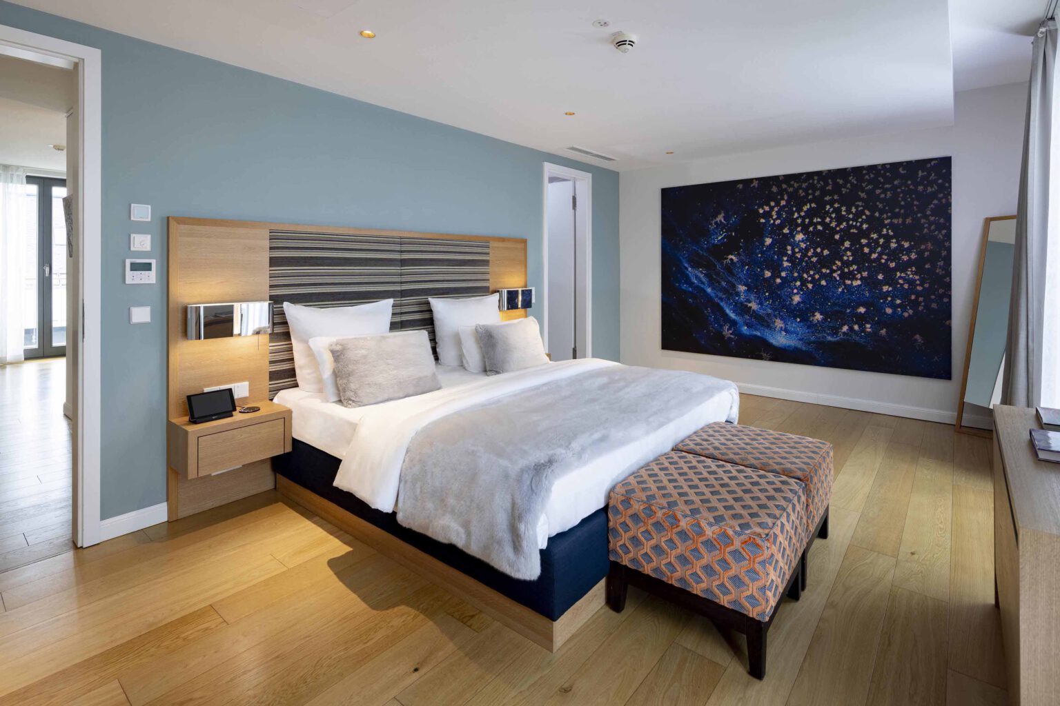 Großzügiges Schlafzimmer der The Wellem Suite mit modernem Kingsize-Bett, eleganter Holzausstattung, hellblauen Wandakzenten und großformatigem Kunstwerk im luxuriösen Hotel The Wellem in Düsseldorf.