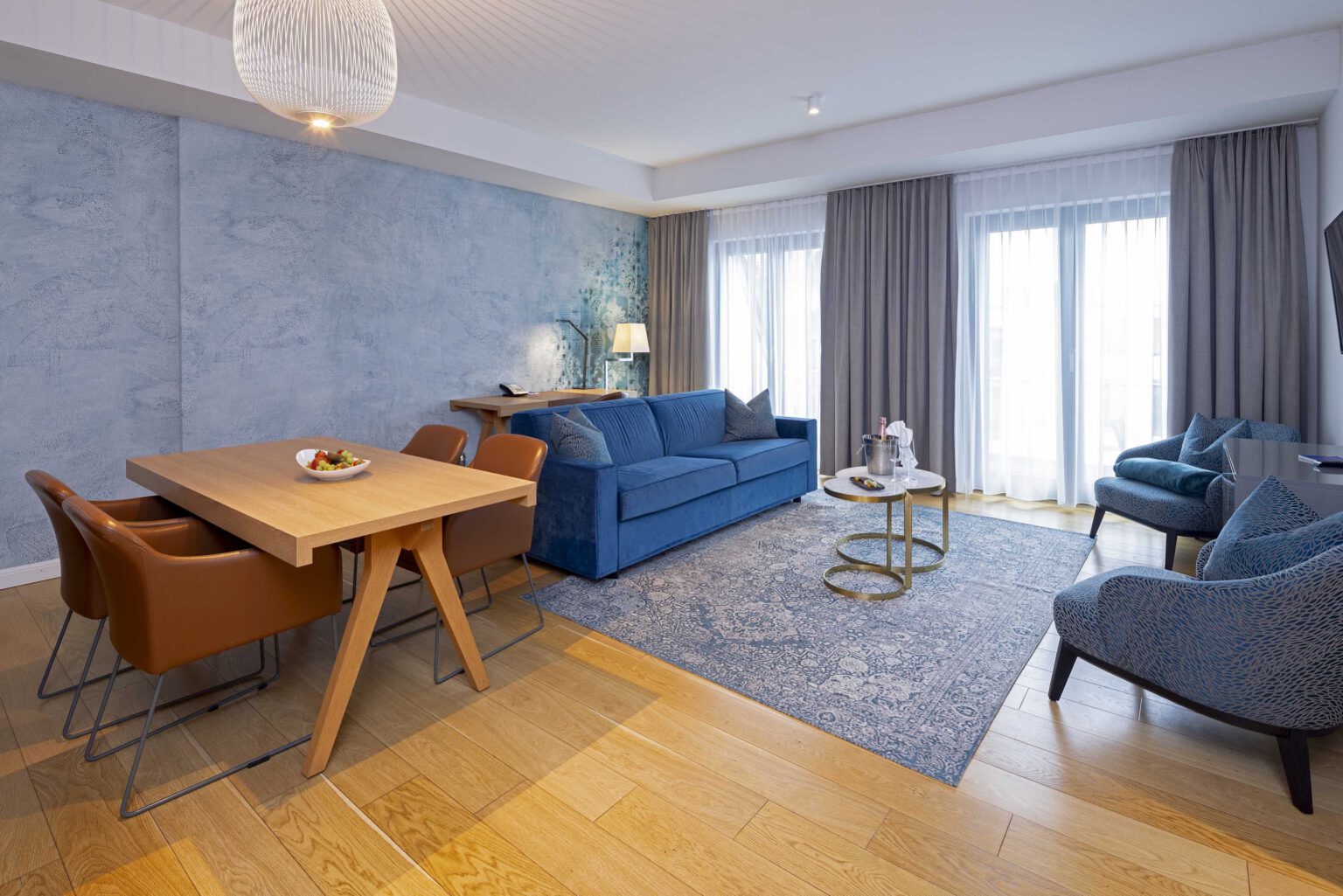 Spacious living area of The Wellem Suite with elegant wooden table, velvet blue couch, designer armchairs, floor-to-ceiling windows and stylish accent wall in the luxurious hotel The Wellem in Düsseldorf.