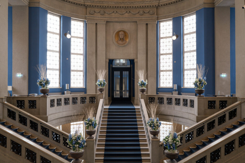 Festlich geschmückte historische Freitreppe in der Eingangshalle des Hotel The Wellem Düsseldorf, umgeben von hohen Fenstern, klassizistischer Architektur und eleganten Blumenarrangements.