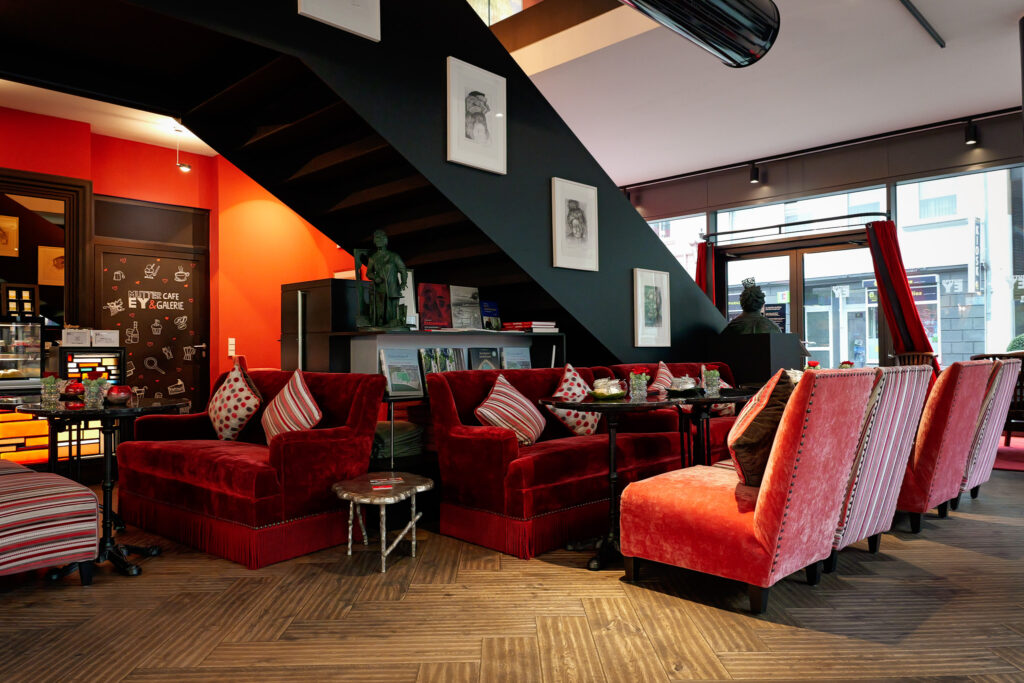 Gemütliche Lounge-Ecke im Mutter Ey Café nahe dem Hotel The Wellem Düsseldorf mit roten Samtsofas, Kunstwerken und warmem Galerie-Ambiente.