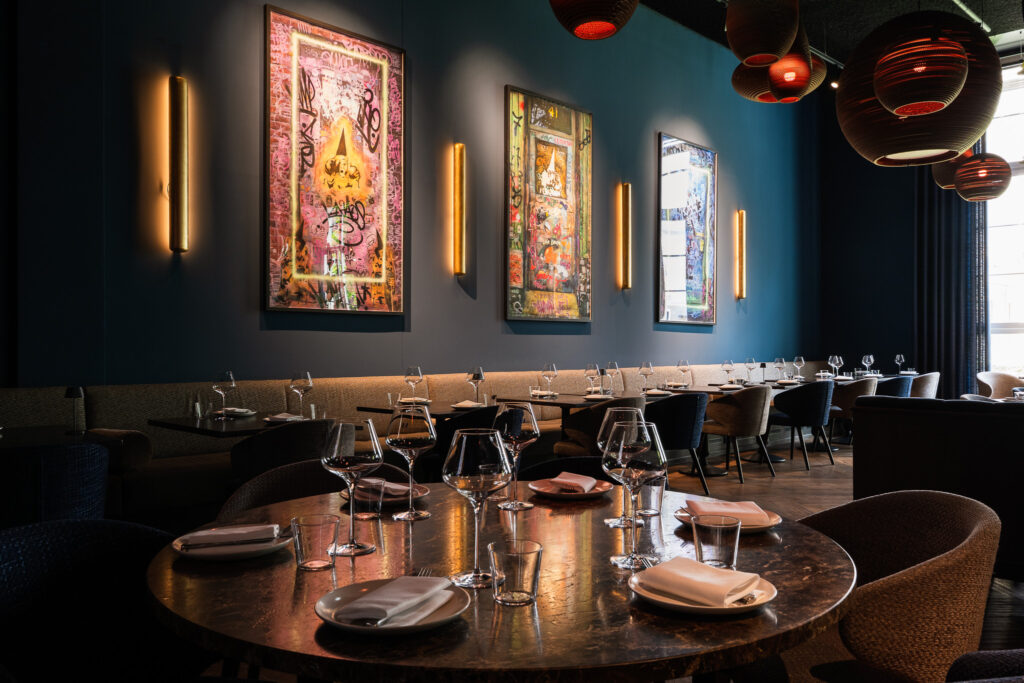 Elegantes Fine-Dining-Ambiente im Restaurant Mbassy by Franks im The Wellem Düsseldorf mit dunklem Interieur, moderner Wandkunst, runden Tischen und eingedeckten Weingläsern.