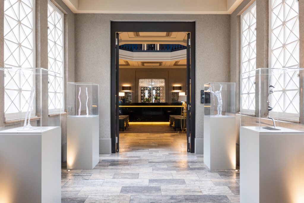 Heller Kunstkorridor im Hotel The Wellem in Düsseldorf mit modernen Skulpturen in Glaskuben, der direkt in die elegante Concierge-Lobby mit Marmortheke und stilvollen Sitzbereichen führt.
