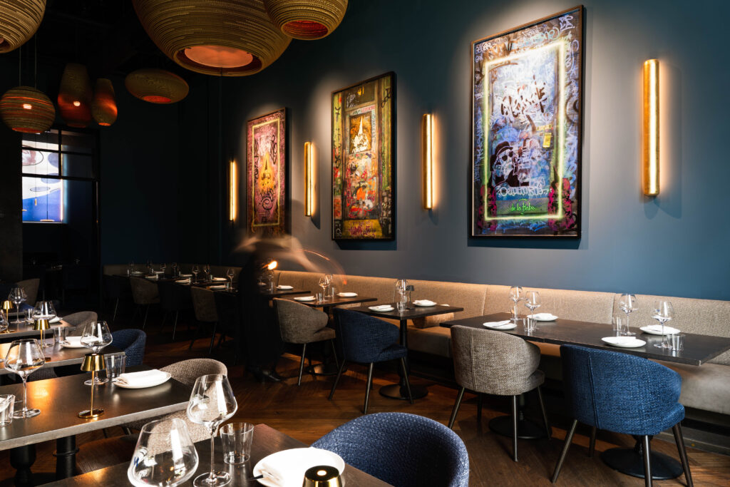 Modernes Fine-Dining-Ambiente im Restaurant Mbassy by Franks im The Wellem Düsseldorf mit farbenfroher urbaner Kunst, eleganter Beleuchtung und stilvoll eingedeckten Tischen.