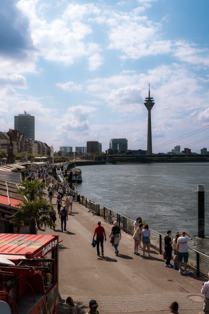Spaziergänger entlang der Rheinuferpromenade mit Blick auf den Rheinturm und die Skyline nahe dem Hotel The Wellem Düsseldorf.